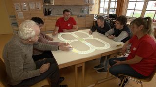 Fünf Personen sitzen um einen Tisch herum und konzentrieren sich auf ein großes beleuchtetes Design einer Eisenbahnstrecke auf dem Tisch. Eine Person zeigt auf das Design.