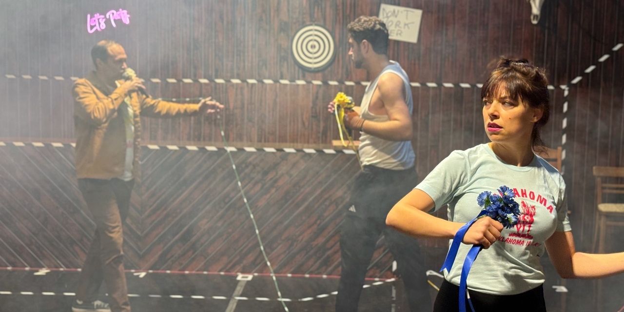 Drei Personen in einem Raum mit einer Holzwand und einem Ziel. Ein Mann mit einer Blume hält ein Band, während ein anderer Mann ein Mikrofon hält und eine andere Frau ein Band hält.