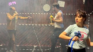 Drei Personen in einem Raum mit einer Holzwand und einem Ziel. Ein Mann mit einer Blume hält ein Band, während ein anderer Mann ein Mikrofon hält und eine andere Frau ein Band hält.