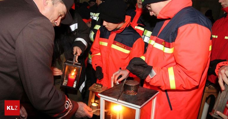 Mehrere Personen in Sicherheitsausrüstung sind um eine Laterne versammelt und zünden Kerzen an. Eine Person mit schwarzem Hut hält eine brennende Kerze, während andere ähnliche Aktivitäten durchführen.