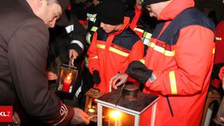 Mehrere Personen in Sicherheitsausrüstung sind um eine Laterne versammelt und zünden Kerzen an. Eine Person mit schwarzem Hut hält eine brennende Kerze, während andere ähnliche Aktivitäten durchführen.