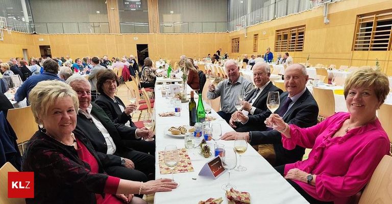 Eine Gruppe älterer Menschen sitzt an einem langen Tisch in einem Saal, hebt Gläser mit Wein, einige lächeln und andere schauen in die Kamera.