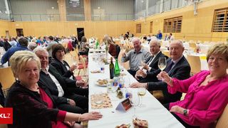 Eine Gruppe älterer Menschen sitzt an einem langen Tisch in einem Saal, hebt Gläser mit Wein, einige lächeln und andere schauen in die Kamera.