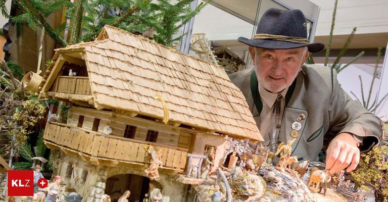 Ein älterer Mann mit Hut steht neben einer detaillierten Holzminiatur eines Dorfhauses mit Figuren und Tieren.