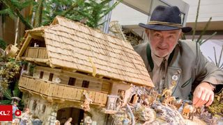 Ein älterer Mann mit Hut steht neben einer detaillierten Holzminiatur eines Dorfhauses mit Figuren und Tieren.
