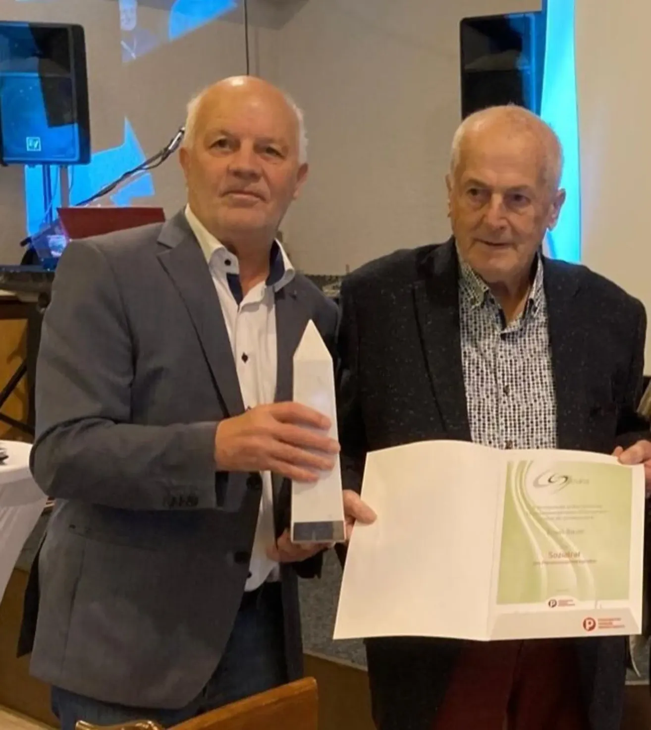 Zwei Männer stehen zusammen, einer hält einen Preis und der andere ein offenes Buch mit einem Zertifikat darin. Sie lächeln beide und posieren für ein Foto.