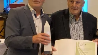 Zwei Männer stehen zusammen, einer hält einen Preis und der andere ein offenes Buch mit einem Zertifikat darin. Sie lächeln beide und posieren für ein Foto.