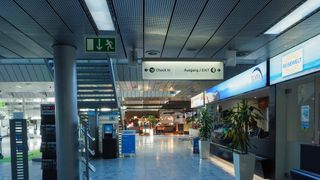 Ein Flughafenterminal mit einem Schild für Check-In und Ausgang/Ausgang. Der Boden ist glänzend und es gibt Topfpflanzen auf beiden Seiten.