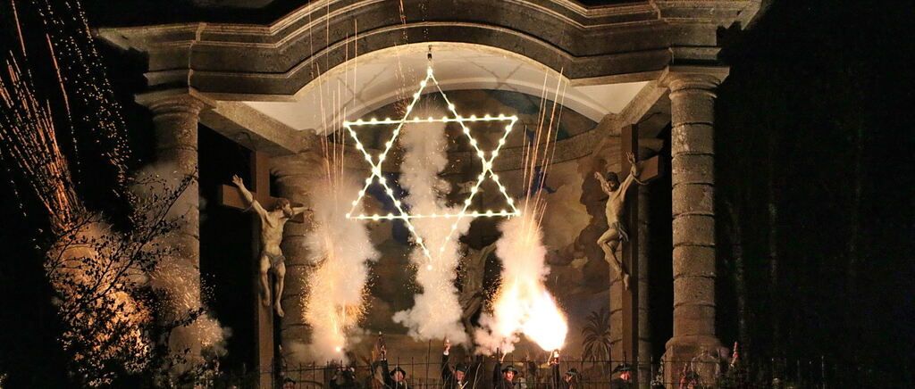 Eine Gruppe von Menschen hält brennende Fackeln und Feuerwerkskörper vor einer sternförmigen Struktur mit Lichtern, nahe einer Wand mit einem Wandbild von Jesus am Kreuz.