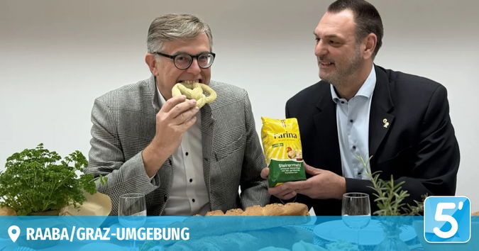 Zwei Männer sitzen an einem Tisch und essen Brezen. Ein Mann hält eine Tüte Brezen und lächelt.