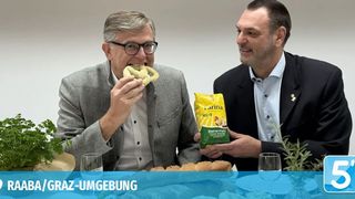 Zwei Männer sitzen an einem Tisch und essen Brezen. Ein Mann hält eine Tüte Brezen und lächelt.