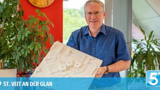 Ein Mann in einem blauen Hemd hält ein großes architektonisches Modell, mit grünen Pflanzen und einer roten Wand im Hintergrund, und einem Logo mit der Nummer 5.