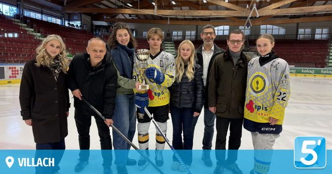 Eine Gruppe von Menschen posiert für ein Foto auf einer Eisbahn, hält Eishockeyschläger und eine Trophäe. Einige tragen Sporttrikots und Winterkleidung. Leere Tribünen und ein Logo sind im Hintergrund zu sehen.