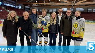 Eine Gruppe von Menschen posiert für ein Foto auf einer Eisbahn, hält Eishockeyschläger und eine Trophäe. Einige tragen Sporttrikots und Winterkleidung. Leere Tribünen und ein Logo sind im Hintergrund zu sehen.
