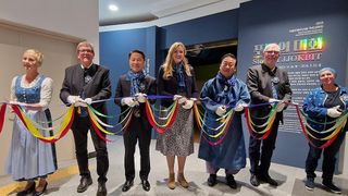 Fünf Personen, drei Männer und zwei Frauen, in formeller Kleidung, schneiden ein Band mit bunten Bändern. Hinter ihnen befindet sich eine blaue Wand mit fremdsprachigem Text.