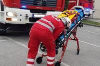Eine Person in roter Notfalluniform schiebt einen Krankenwagen mit einem Patienten im Freien. Ein Feuerwehrwagen ist hinter ihnen geparkt.