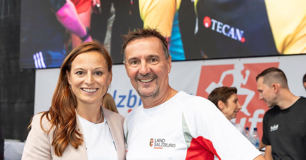 Ein Mann und eine Frau stehen lächelnd bei einer Veranstaltung, tragen weiße T-Shirts mit Logos für Land Salzburg. Dahinter steht eine verschwommene Figur und eine große Leinwand mit einem Bild eines Läufers.