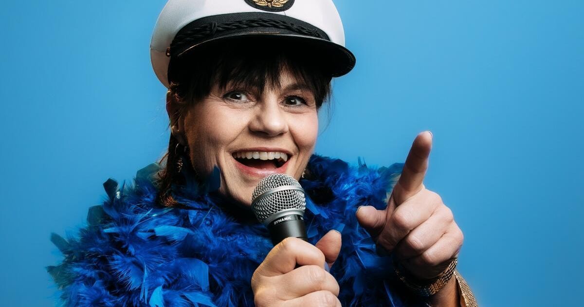 Eine Frau in einer weißen Mütze mit einem blauen Federboa hält ein Mikrofon, lächelt und zeigt mit dem Zeigefinger vor einem blauen Hintergrund.