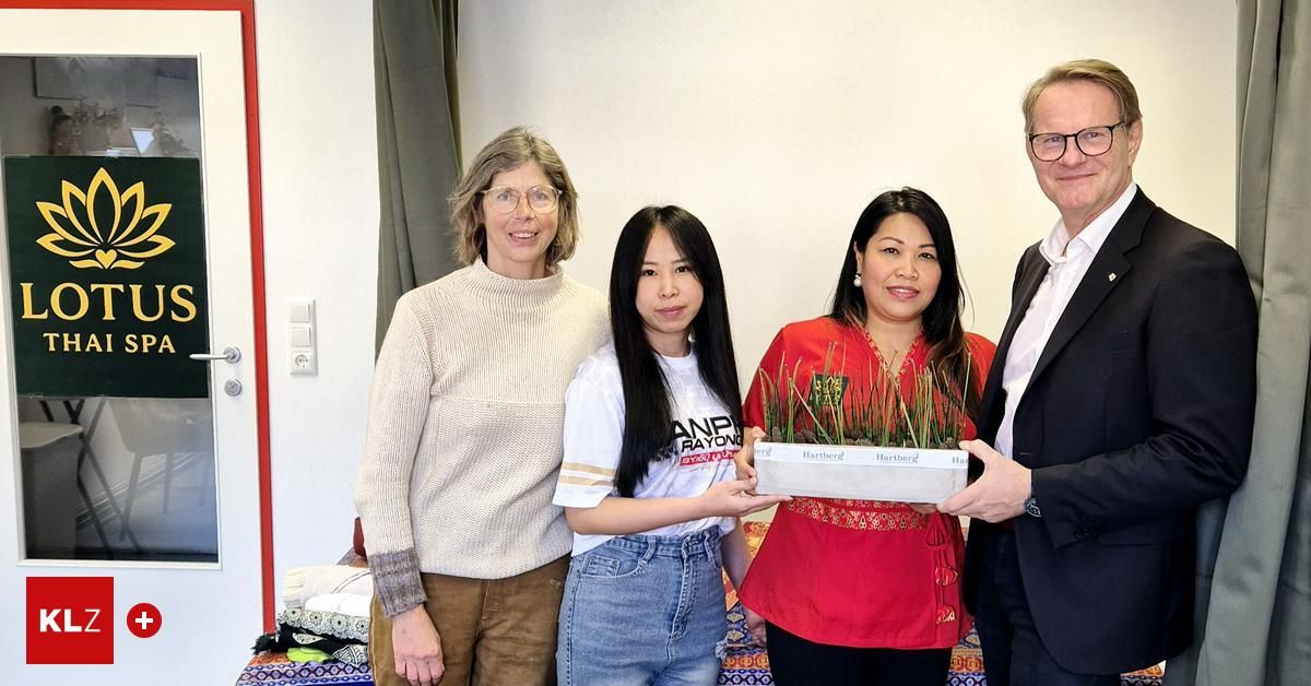 Vier Menschen stehen in einem Raum mit einem Pflanzenkasten und lächeln. Eine Frau in Rot hält den Kasten und eine andere in Weiß trägt ein T-Shirt. Ein Mann in einem Anzug steht neben ihnen. Hinter ihnen hat eine weiße Wand eine Steckdose und einen Vorhang.