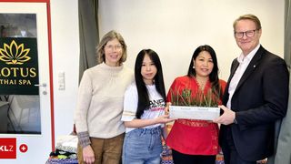 Vier Menschen stehen in einem Raum mit einem Pflanzenkasten und lächeln. Eine Frau in Rot hält den Kasten und eine andere in Weiß trägt ein T-Shirt. Ein Mann in einem Anzug steht neben ihnen. Hinter ihnen hat eine weiße Wand eine Steckdose und einen Vorhang.