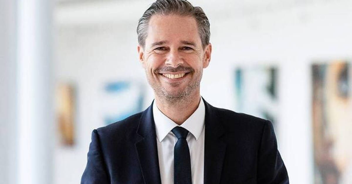 Ein lächelnder Mann in Anzug und Krawatte, der in einem modernen Büro steht. Er hat kurze graue Haare und einen Bart.