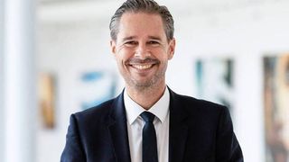 Ein lächelnder Mann in Anzug und Krawatte, der in einem modernen Büro steht. Er hat kurze graue Haare und einen Bart.