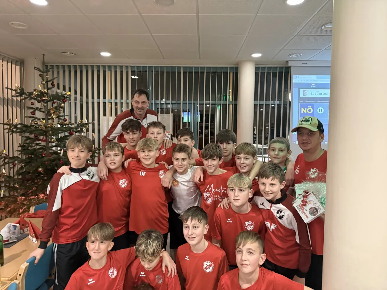Eine Gruppe junger Jungen in roten Trikots, wahrscheinlich eine Fußballmannschaft, posiert für ein Foto mit ihrem Trainer in einem Raum mit einem Weihnachtsbaum und Jalousien.