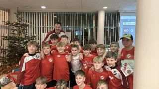 Eine Gruppe junger Jungen in roten Trikots, wahrscheinlich eine Fußballmannschaft, posiert für ein Foto mit ihrem Trainer in einem Raum mit einem Weihnachtsbaum und Jalousien.