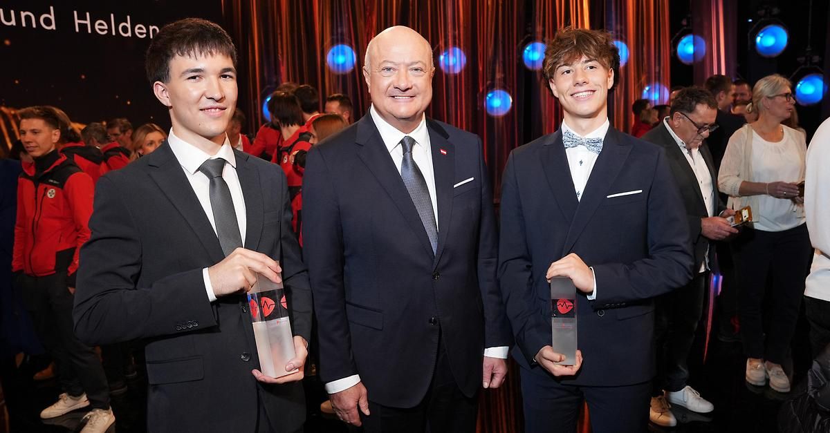 Drei Personen stehen zusammen und halten Auszeichnungen. Der Mann in der Mitte lächelt. Die beiden Jungen auf beiden Seiten halten Auszeichnungen. Hinter ihnen ist ein roter Vorhang mit blauen Lichtern zu sehen.