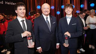 Drei Personen stehen zusammen und halten Auszeichnungen. Der Mann in der Mitte lächelt. Die beiden Jungen auf beiden Seiten halten Auszeichnungen. Hinter ihnen ist ein roter Vorhang mit blauen Lichtern zu sehen.