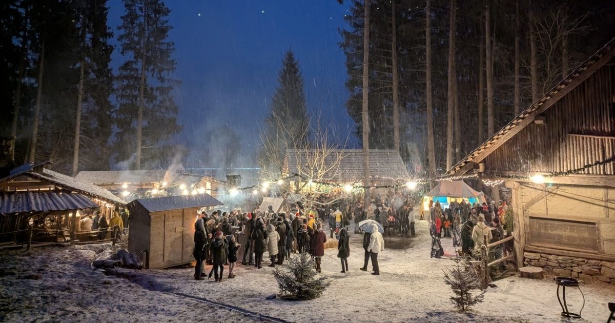 Ein schneebedeckter Outdoor-Markt bei Nacht, mit Menschen in Winterkleidung. Mehrere Holzstrukturen mit Lichtern und Dekorationen. Bäume im Hintergrund sind mit Schnee bedeckt.