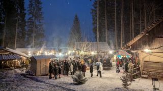 Ein schneebedeckter Outdoor-Markt bei Nacht, mit Menschen in Winterkleidung. Mehrere Holzstrukturen mit Lichtern und Dekorationen. Bäume im Hintergrund sind mit Schnee bedeckt.