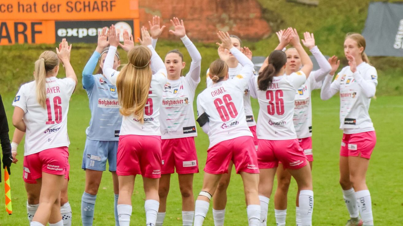 Eine Gruppe von Fußballspielerinnen in pinkfarbenen Trikots, wahrscheinlich nach einem Sieg, heben ihre Hände auf einem Rasenfeld. Das Wort 'GRAFL' und die Zahlen '66' sind auf einem Spielertrikot zu sehen.