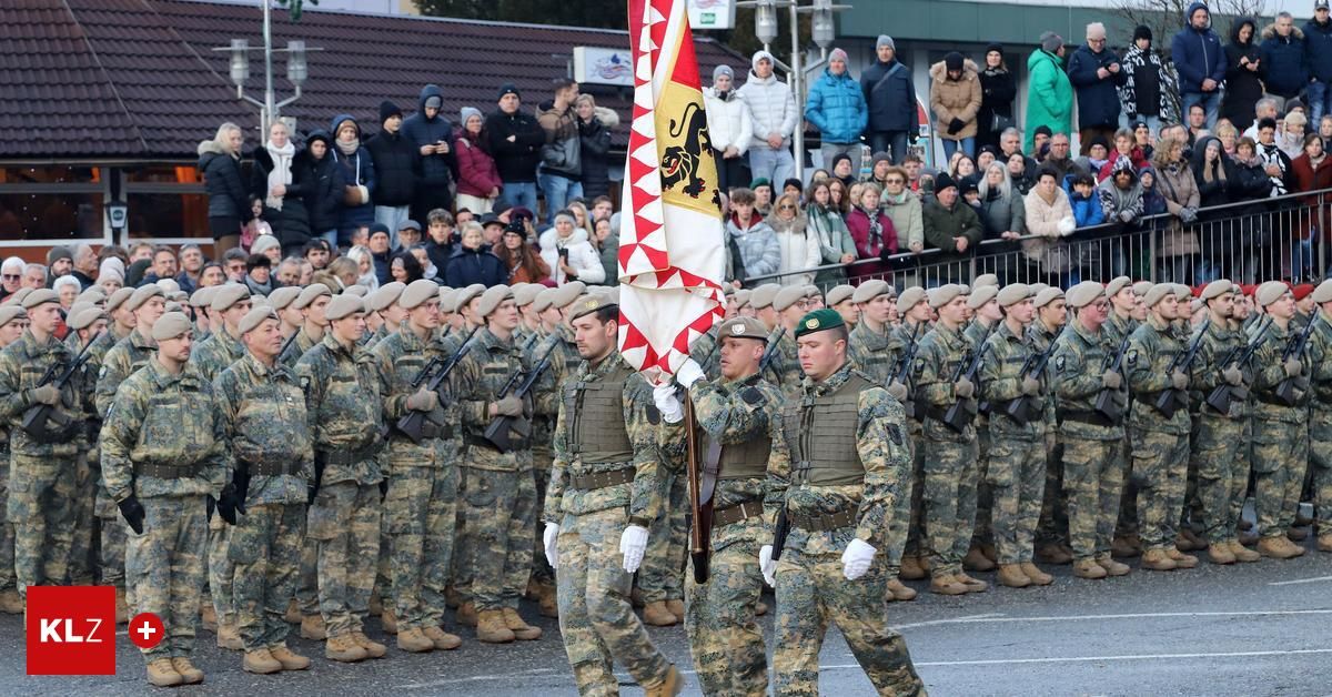Ein Militärparade mit Soldaten, die Gewehre und eine Flagge tragen, marschiert vor einer Menge.