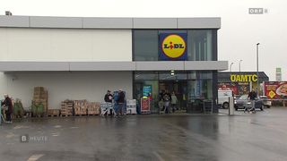 Menschen kaufen am Eingang eines Lidl-Geschäfts ein. Mehrere Personen schieben Einkaufswagen. Ein weißer Lieferwagen ist draußen geparkt.