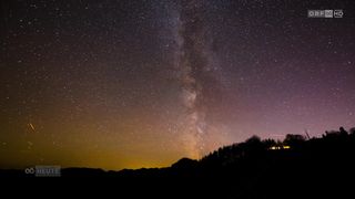 Der Nachthimmel ist voller Sterne und die Milchstraße ist sichtbar und taucht die Silhouette von Bäumen und Bergen in ein sanftes Licht.