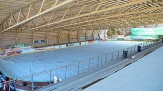 Eine leere Eishalle für Eishockey mit Metallgeländern und einem Mülleimer. Die Eisfläche hat blaue Markierungen und ist von Metallkonstruktionen umgeben.