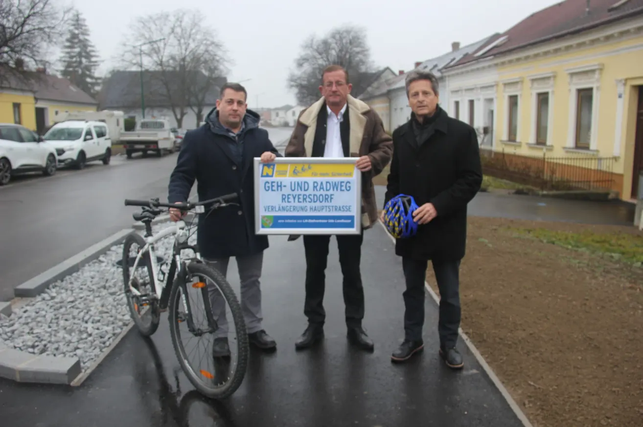Drei Männer posieren auf einem Bürgersteig mit einem Schild, das Geh-und Radweg Reyersdorf besagt. Der Mann auf der linken Seite hält ein Fahrrad, während die anderen zwei Helme halten.