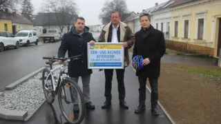 Drei Männer posieren auf einem Bürgersteig mit einem Schild, das Geh-und Radweg Reyersdorf besagt. Der Mann auf der linken Seite hält ein Fahrrad, während die anderen zwei Helme halten.