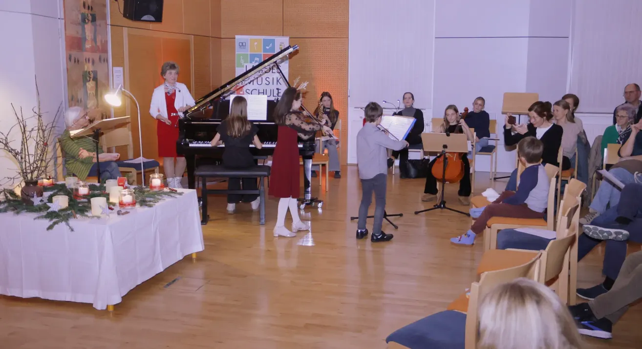 Eine Gruppe von Musikern tritt in einem Saal auf. Eine Frau in einem roten Kleid spielt Klavier, während eine andere Frau Cello spielt. Ein Junge spielt Trompete, und andere Menschen spielen verschiedene Instrumente. Ein Tisch mit weißer Tischdecke und einer Kerze steht vor ihnen.