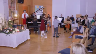 Eine Gruppe von Musikern tritt in einem Saal auf. Eine Frau in einem roten Kleid spielt Klavier, während eine andere Frau Cello spielt. Ein Junge spielt Trompete, und andere Menschen spielen verschiedene Instrumente. Ein Tisch mit weißer Tischdecke und einer Kerze steht vor ihnen.