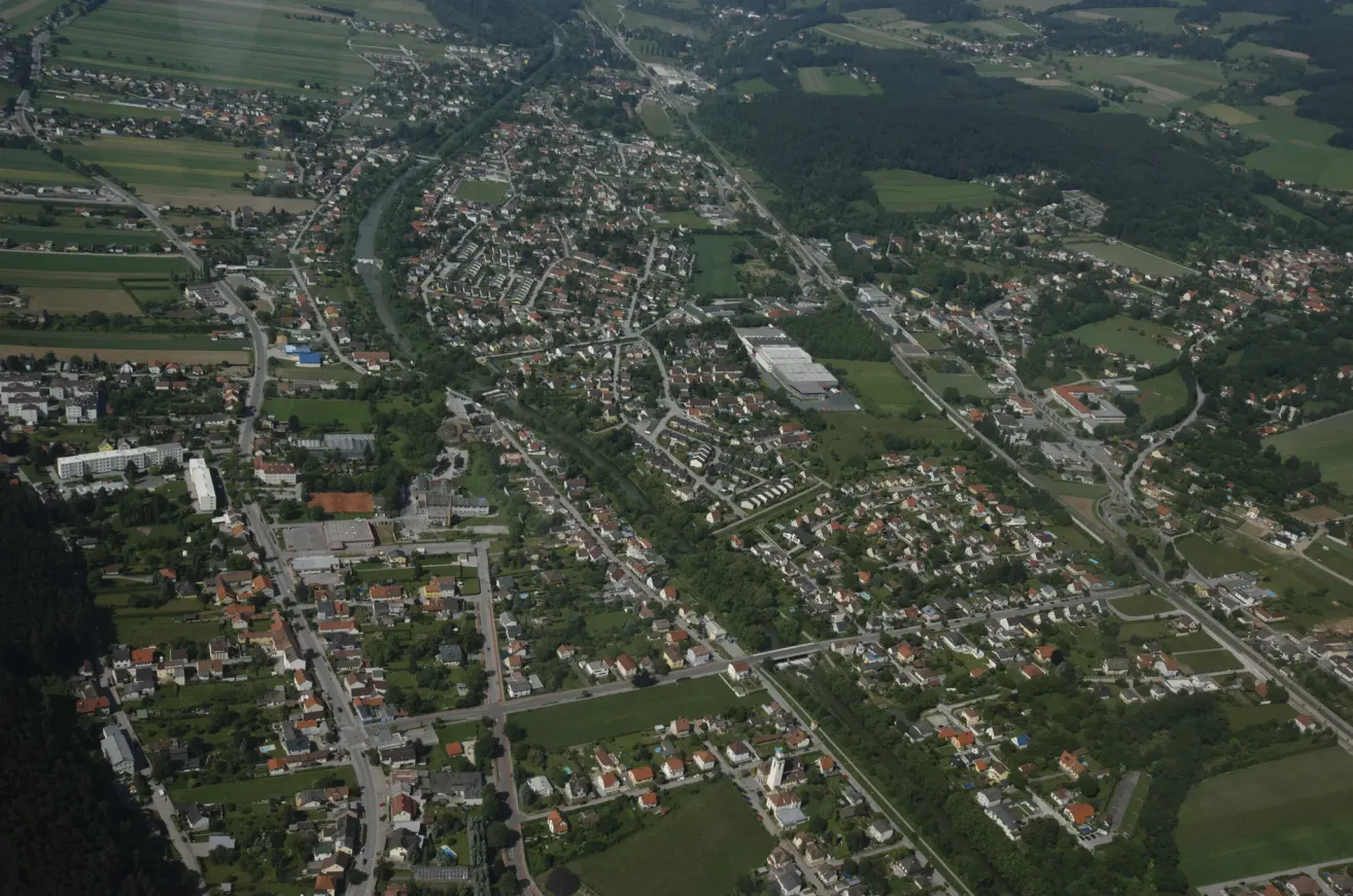Luftaufnahme einer Stadt mit vielen Gebäuden, Straßen und Grünflächen. Die Stadt ist von Bäumen und Vegetation umgeben, durch die ein Fluss fließt.