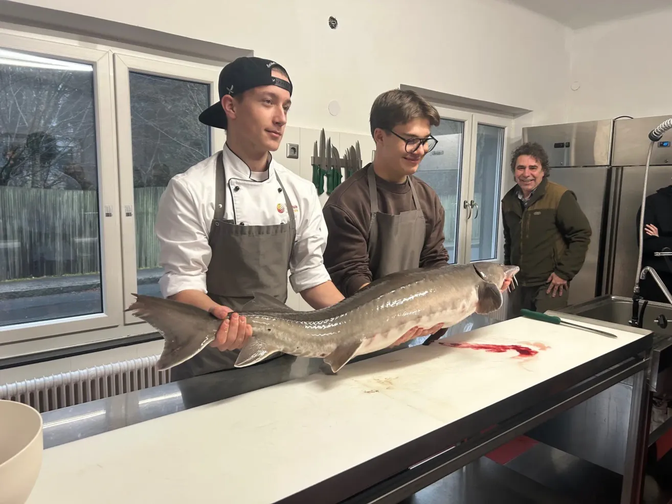 Drei Personen stehen in einer Küche. Zwei von ihnen halten einen großen Fisch, während eine dritte Person hinter ihnen lächelt.