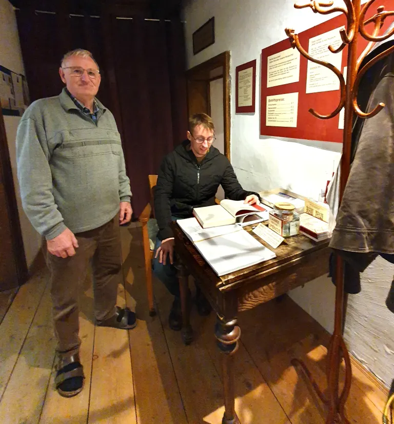 Zwei Personen stehen in einem Raum. Eine sitzt an einem Schreibtisch mit Büchern, während die andere in der Nähe steht. Ein Kleiderständer und eine Pinnwand befinden sich an der Wand.
