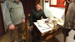 Zwei Personen stehen in einem Raum. Eine sitzt an einem Schreibtisch mit Büchern, während die andere in der Nähe steht. Ein Kleiderständer und eine Pinnwand befinden sich an der Wand.