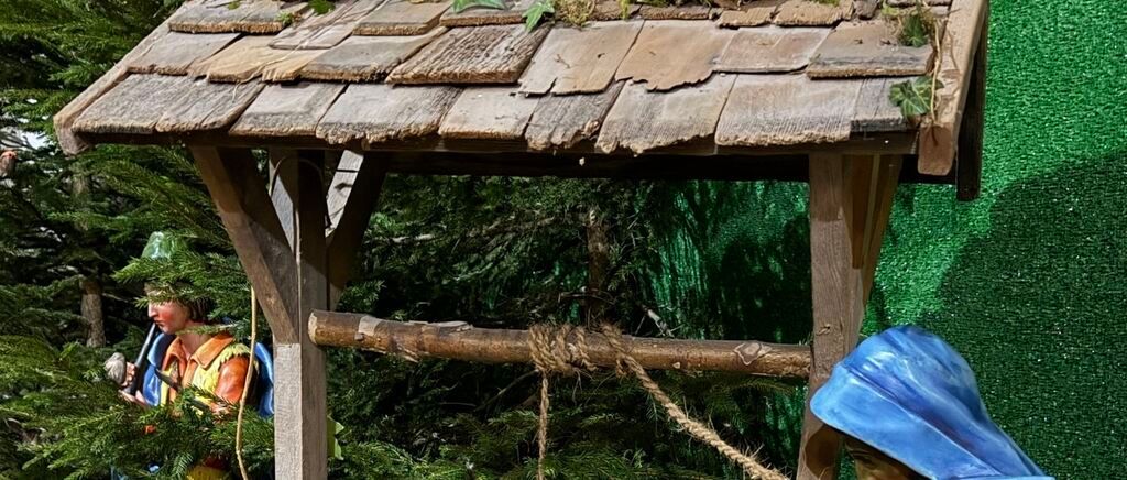 Eine Krippe mit einer Holzstruktur, einem blauen Hut und einem Seil. Zwei Figuren sind vorhanden, eine trägt ein gelbes Shirt und die andere ein blaues Weste.