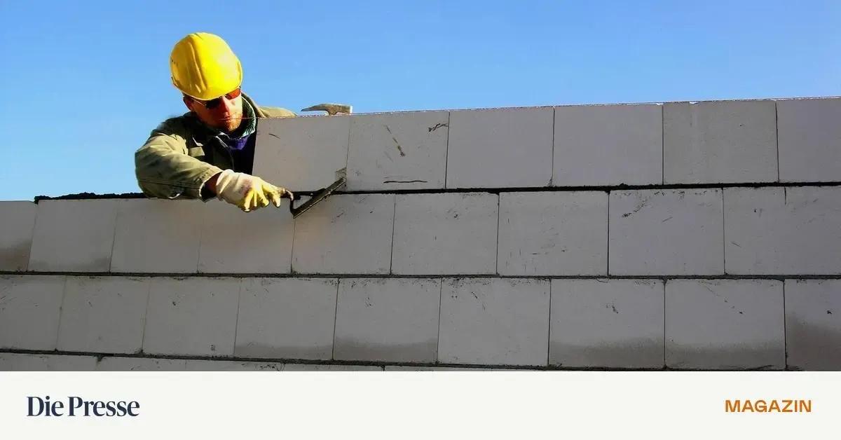 Ein Mann mit gelbem Schutzhelm und Handschuhen benutzt eine Kelle, um Zement auf eine weiße Ziegelmauer aufzutragen.