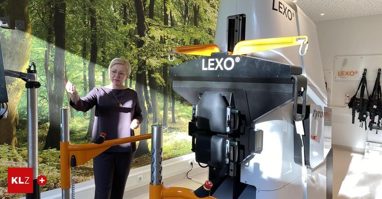 Eine Frau steht neben einer großen, gelb-schwarzen Maschine mit dem Wort 'LEXO' darauf und zeigt darauf. Der Hintergrund ist ein Wandbild eines Waldes.