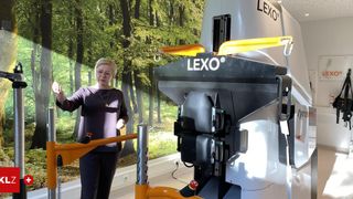 Eine Frau steht neben einer großen, gelb-schwarzen Maschine mit dem Wort 'LEXO' darauf und zeigt darauf. Der Hintergrund ist ein Wandbild eines Waldes.
