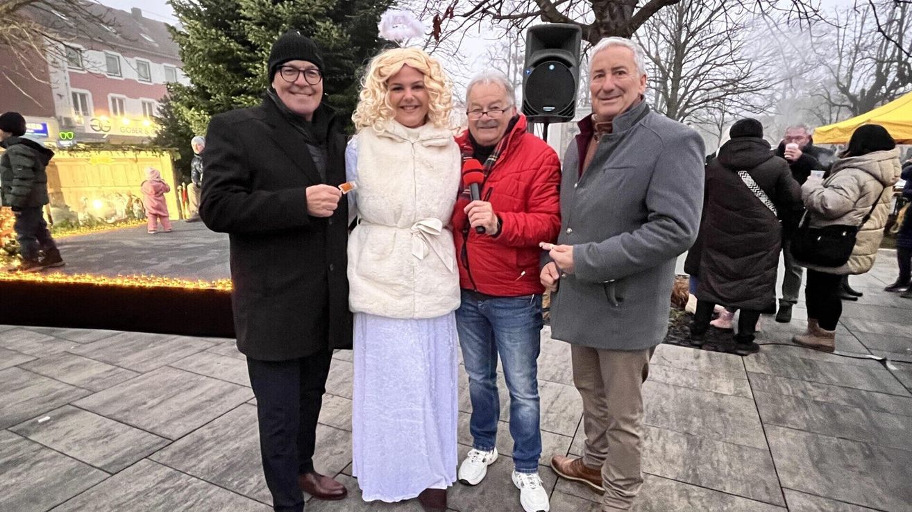 Vier Personen stehen in einem Park und lächeln für ein Foto. Eine Frau in einem weißen Kleid steht zwischen zwei Männern, einer hält ein Mikrofon. Hinter ihnen steht ein Lautsprecher und Bäume.